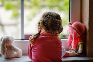 Cute toddler girl sitting by window and looking out on rainy day. Dreaming child with doll and toy...