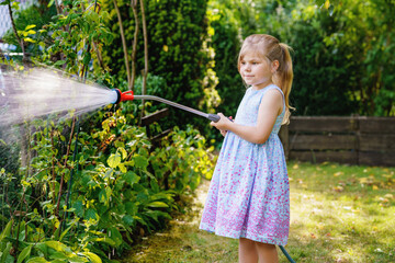 Happy little girl playing with a garden hose on hot and sunny summer evening. Preschool child helping and having fun with watering trees and plants in domestic garden