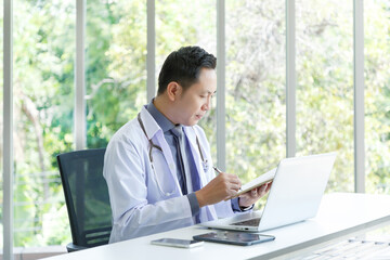 Asian senior doctor sitting in medical office while taking note and using laptop computer.