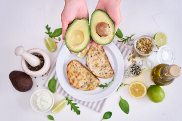 Avocado and cream cheese toasts preparation - Woman holding halved avocado over table with ingredients