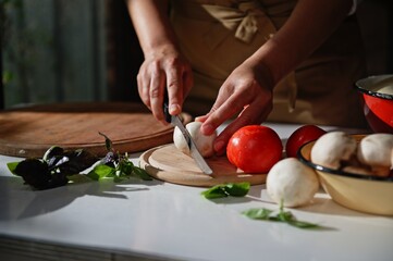 Hands of a chef chopping fresh mushroom champignons on a wooden cutting board near organic vegetables and culinary herbs