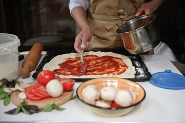 Front view housewife in beige chef apron spreads freshly made tomato sauce on rolled out dough. Italian cuisine Culinary