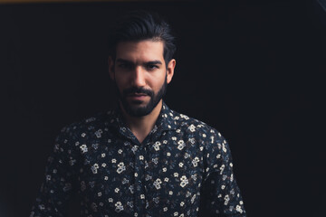Obraz premium Hispanic man with dark hair and beard wearing dark floral shirt looking into camera with intimidating expression. Dark background studio shot. High quality photo