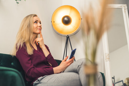 Blonde white woman wearing maroon shirt in living room putting pinger to chin thinking and looking up holding smartphone. Indoor shot. High quality photo