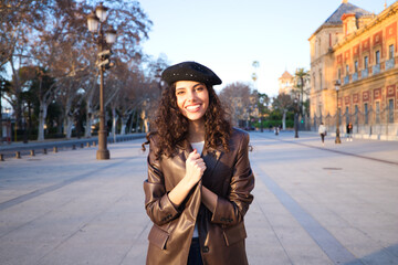 Pretty young woman with curly brown hair wearing a black cap and dressed in a leather jacket, jeans and white boots. She is sightseeing in the city. Concept of holidays and tourism around the world.
