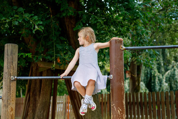 Little preschool girl climbing and hanging on outdoor horizontal bar. Cheerful girl athlete on gymnastic stick on sports ground. Happy child and summer activities on playground.