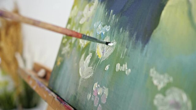 Person hand using paintbrush paints white flowers on green land on picture canvas. Skilled master creates masterpiece painting watercolor landscape closeup
