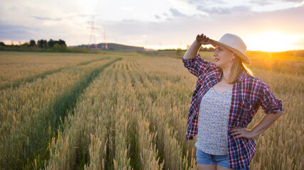 Fototapeta na wymiar A beautiful middle-aged farmer woman in a straw hat and a plaid shirt stands in a field of golden ripening wheat during the daytime in the sunlight