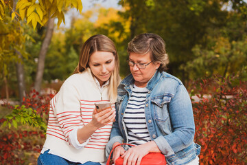 charming women in casual style, mom and daughter are sitting on bench in beautiful autumn park and looking at photos on phone - obrazy, fototapety, plakaty