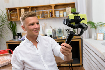 Blond laughing guy filming a video blog on a mobile device using an electronic image stabilizer in the kitchen - obrazy, fototapety, plakaty