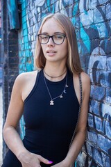 a beautiful blonde girl in glasses and a black dress stands against a wall with graffiti