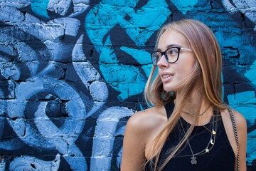 a beautiful blonde girl in glasses and a black dress stands against a wall with graffiti