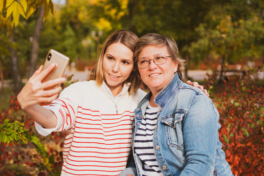 adorable women , mom and daughter are sitting on bench in beautiful autumn park and taking selfie on phone