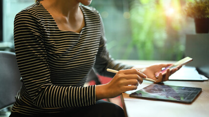 Young creative woman holding holding mobile phone and using digital tablet at her workplace