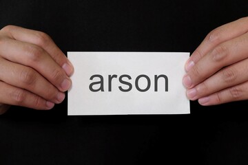 Arson crime case mug shot concept. Criminal hands holding paper placard with written word in dark black background.