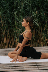 Eine junge tätowierte Frau macht Yoga auf einem Steg am See. Yoga Position pigon post. 1 Yoga und Meditation am See 1