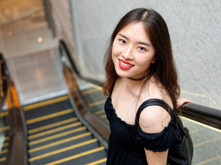 Portrait of beautiful Chinese young girl wearing black tube dress standing on elevator and smiling at camera in shopping mall, indoor fashion portrait of glamour sexy stylish lady with black long hair