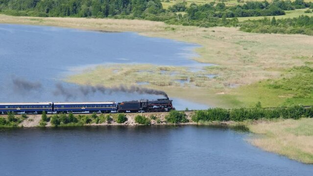 Russia Republic Karelia. Popular tourist destination. Ruskeala express retro train crosses river bridge black smoke billows from chimney. View from Mount Paasonvuori in Sortavala to lake and fields.