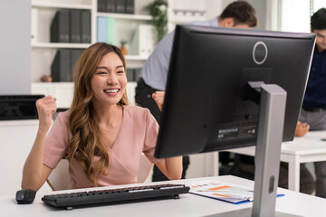 Asian female employee raising hands happy and laugh for receiving good news or project successful looking on computer at desk in the office. Salary growth or career promotion. Success work