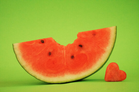 St&uuml;ck Wassermelone vor gr&uuml;nem Hintergrund, horizontal, in der Mitte angebissen und ausgestochenes Herz aus Melone daneben 