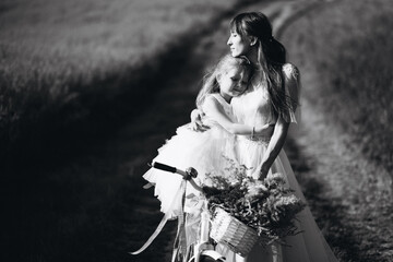 Mother with little daughter wearing beautiful dresses riding bicycle in the field
