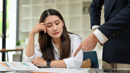 A male boss complains to a tired and bored young Asian female accountant.