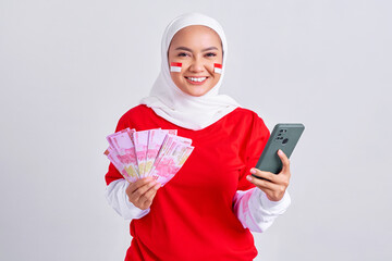 Smiling young Asian muslim woman in red white t-shirt holding mobile phone and cash money in Indonesian rupiah banknotes isolated on white background. Indonesian independence day on 17 august concept