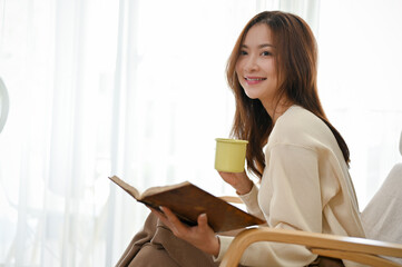 Attractive young Asian female enjoys reading a book while sipping coffee in her living room.