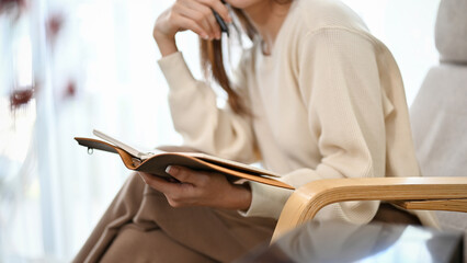 Relaxed and calm young Asian female sits on comfy armchair reading a book. cropped image