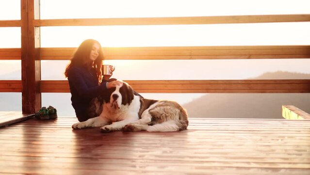 Beautiful woman enjoying cup of coffee or tea on warm sunset in the mountains and playing with her saint bernard dog.