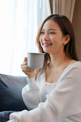 Young Asian female is chilling and relaxing in the living room, having a morning coffee.