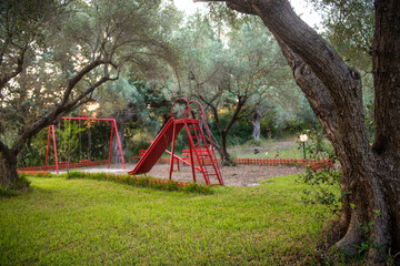 Fototapeta na wymiar Kids playground under the olive trees in forest. Childrens play area in the park. Safe beautiful red natural playground for children in forest.
