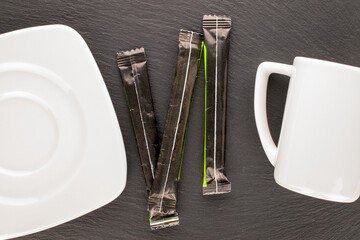 Three sticks of sugar in paper bags with a white saucer and a cup on a slate stone, close-up, top...