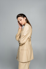 brunette model in beige blazer looking at camera while posing isolated on grey