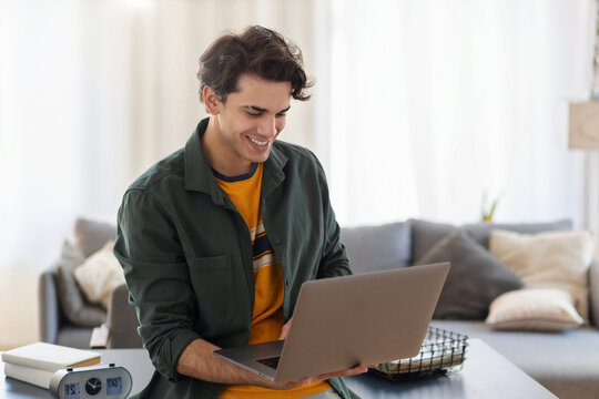 Young man freelancer using laptop for work while standing near workplace in a modern office or home, online remote work, smiling