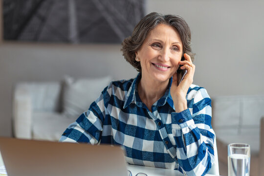 Beautiful senior woman working talking on mobile phone sitting at the workplace with computer laptop in a modern office or home, online remote work, smiling