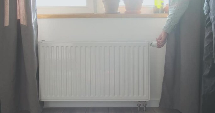 Unrecognizable man turns off the thermostat on the radiator near the window. Bright sunlight comes through the window. The concept of spring, warm weather, heating savings.