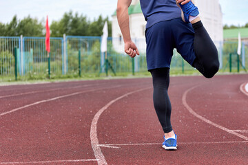 Obraz premium Runner kneads his legs in stadium before race on a circular treadmill with a rubber coating. Long-legged athlete in a sports uniform prepares for the starting race, kneads the muscles of the legs