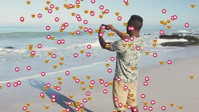 Animation of heart and love emojis over african american man taking smartphone photos on sunny beach