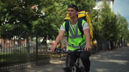 Unsure courier in green vest driving bicycle looking around in residential neighborhood. Portrait of young Caucasian delivery man riding bike delivering order in urban city