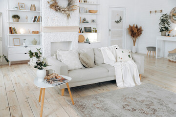 Scandinavian style sofa with plaid and pillows and coffee table in modern stylish bright living room with fireplace, shelving and console table. Nobody