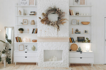 White shelving with books, decor and potted plants near fireplace with wreath of dried flowers in modern stylish living room in scandinavian style. Nobody