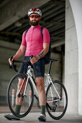 Naklejka premium Portrait of a cyclist standing under a bridge with a bicycle in his hand, posing at the camera against an architecture background. Active lifestyle. Cycling is a hobby.