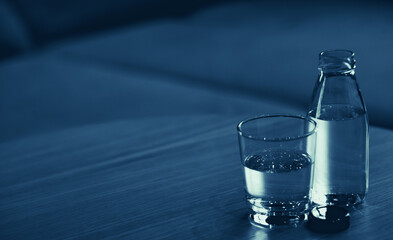 Glass and bottle of drinking water on table in empty room, sofa with pillows in background.