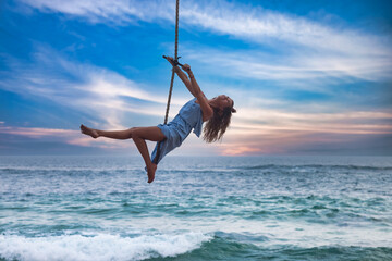 Young slim woman in blue dress swing on bungee, flying over ocean sunset