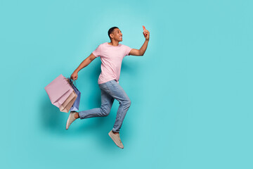 Full length photo of cheerful positive guy wear pink t-shirt jumping holding bags selfie empty space isolated teal color background
