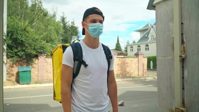 Positive Delivery Boy In Coronavirus Face Mask Passing Parcel To Client At Gate In Town. Portrait Of Caucasian Young Man Delivering Food And Drink On Covid-19 Pandemic Outbreak