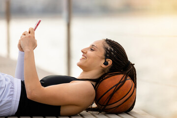 Athletic young woman smiling happy wearing wireless smartphone earphones lying on basketball