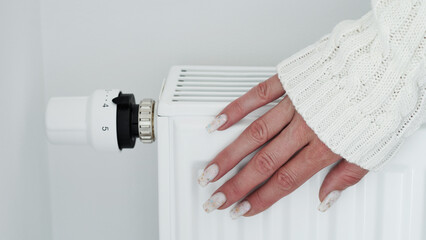 Close-up of a woman in white sweater warms her hands over the battery on background of white wall.Macro photo of woman's hands,copy space.Concept of saving gas in winter.Gas crisis in Europe.