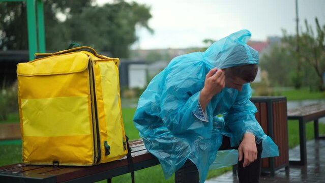 Exhausted young courier sitting in rain coat on bench in city park with yellow backpack. Portrait of stressed tired Caucasian delivery man waiting orders outdoors on rainy spring day